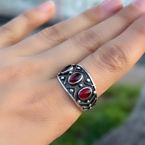 Op de afbeelding: Een zilveren ring met drie rode ovale edelstenen, gezet in een gedetailleerd ontwerp.