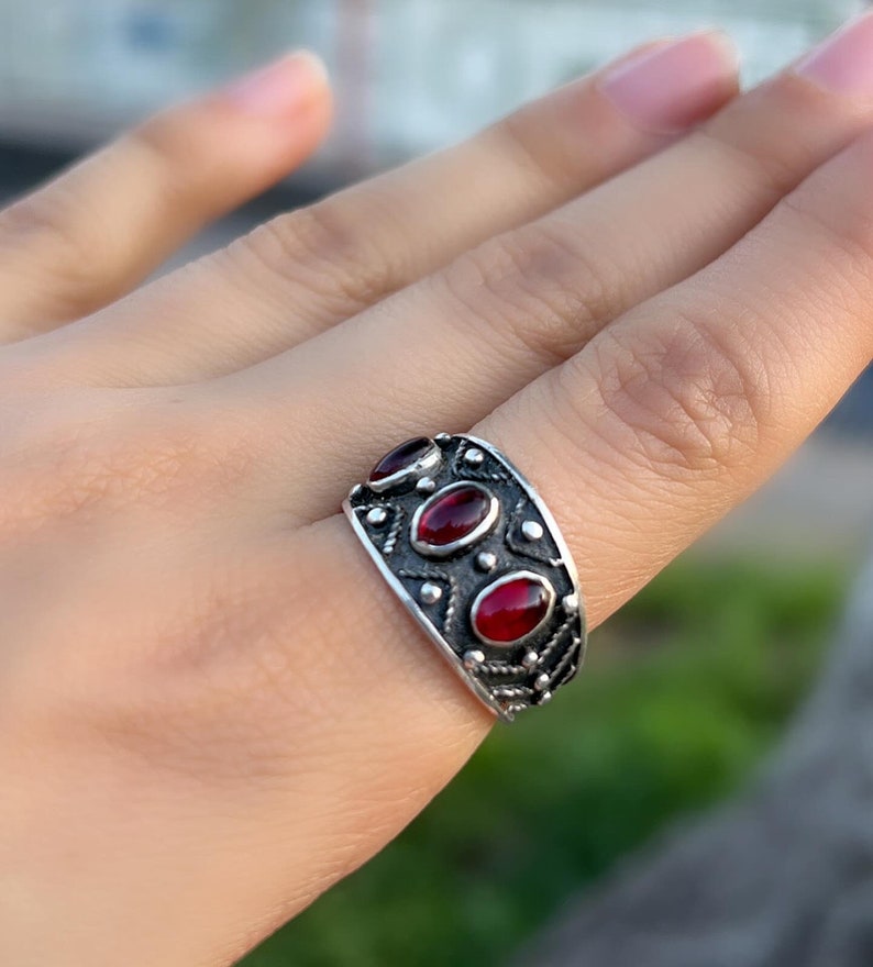Op de afbeelding: Een zilveren ring met drie rode ovale edelstenen, gezet in een gedetailleerd ontwerp.