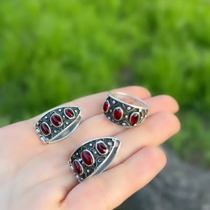 Op de afbeelding: Een set van drie zilveren ringen met rode edelsteen accenten. De ringen zijn in de vorm van een band en een traan. De ringen liggen op een hand tegen een groene achtergrond.