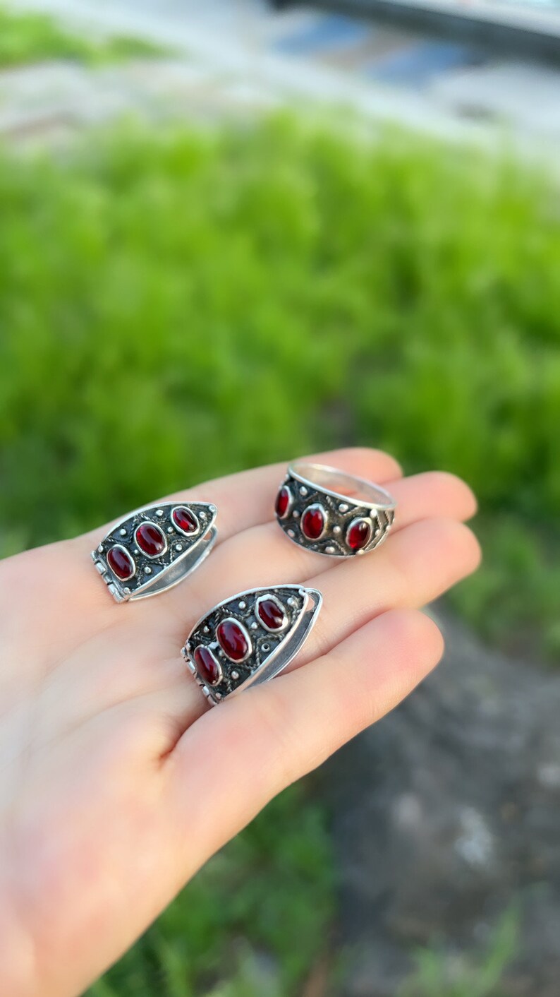 Op de afbeelding: Een set van drie zilveren ringen met rode edelsteen accenten. De ringen zijn in de vorm van een band en een traan. De ringen liggen op een hand tegen een groene achtergrond.