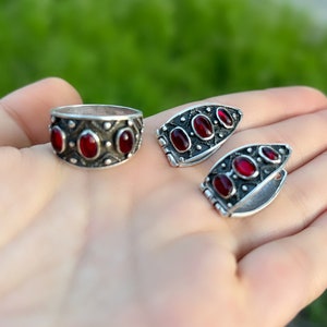 Op de afbeelding: Een set zilveren ringen met rode edelstenen. De ring is een band met drie ovale stenen. De oorbellen zijn een paar gebogen hoepels met drie ovale stenen.