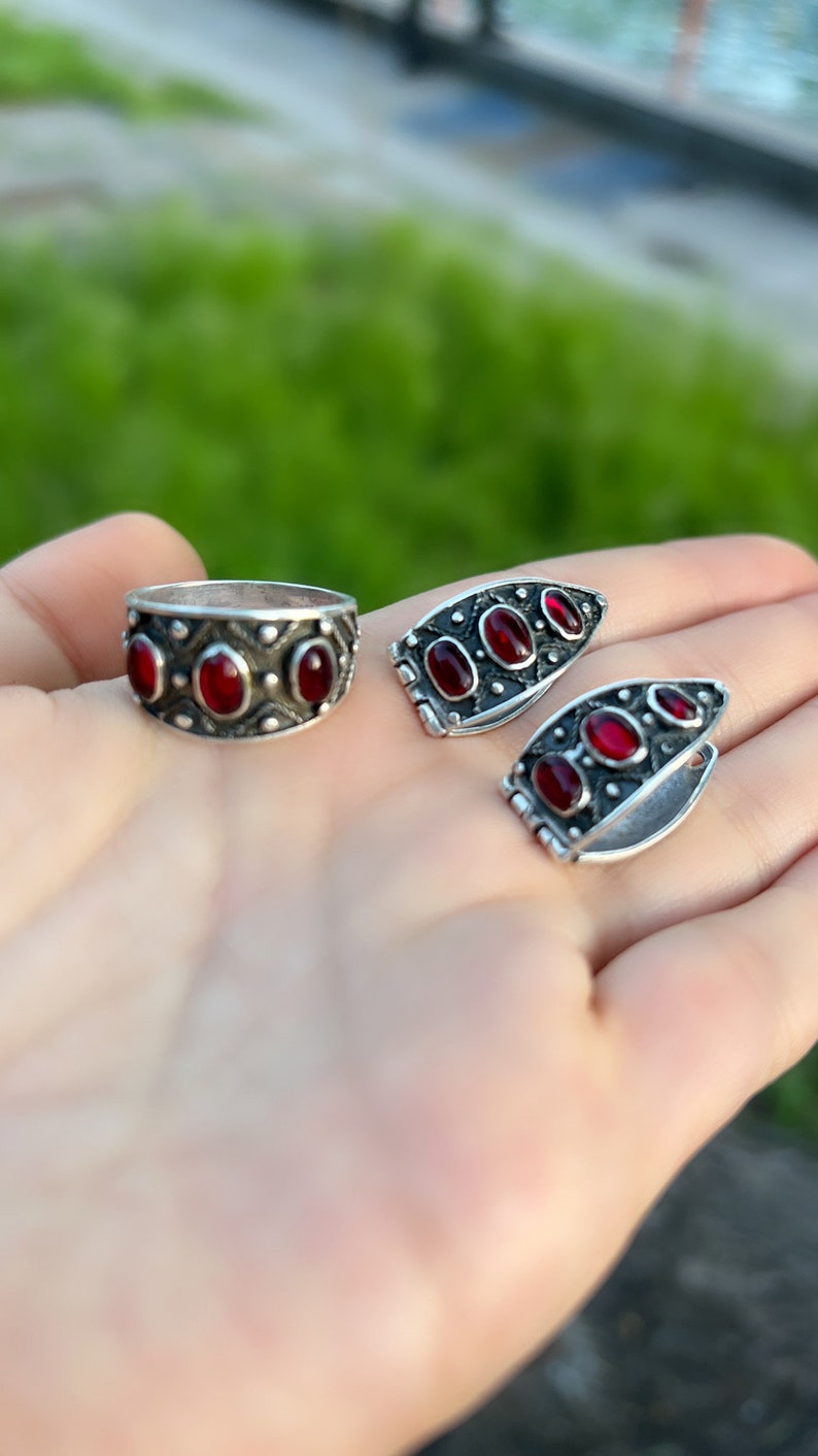 Op de afbeelding: Een set zilveren ringen met rode edelstenen. De ring is een band met drie ovale stenen. De oorbellen zijn een paar gebogen hoepels met drie ovale stenen.