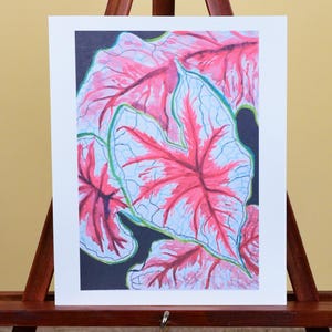 May include: A rectangular art print featuring a botanical illustration of heart-shaped leaves with white centers, vibrant pink veins, and green edges set against a solid black background, displayed on a dark brown wooden tabletop easel.