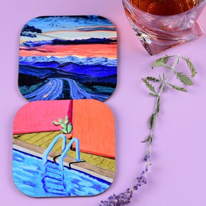 May include: Two square coasters with colorful landscape paintings, one of a road through mountains at sunset, the other of a pool with a cactus. A glass of amber liquid and sprig of lavender are also present.