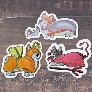 May include: Three illustrated stickers of corgis dressed up in costumes. One corgi is dressed as a vampire, another as an angel, and the last as a devil.