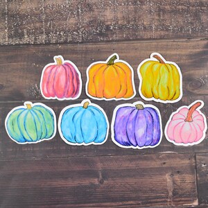 May include: Seven hand-painted pumpkin stickers in various colors, including pink, orange, yellow, green, blue, purple, and light pink. The stickers are arranged in a semi-circle on a brown wooden background.