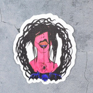 May include: A sticker featuring a black and white illustration of a woman with long, dark hair. Her face is pink and she has a spider on her chest. The background is a gray, textured surface.