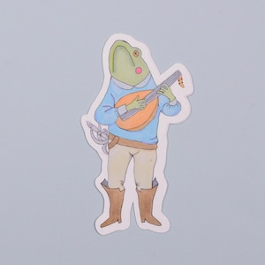May include: A green frog wearing a blue shirt and brown pants plays a lute with a yellow body. The frog has a red mouth and is wearing brown boots.