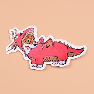 May include: A sticker featuring a dog in a red dragon costume. The dragon costume has brown wings and a tail. The dog's face is visible, with brown and white fur. The sticker is on a peach-colored background.