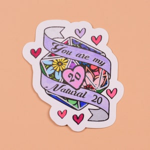May include: A purple and white sticker with a colorful illustration of a 20-sided die. The die has a heart in the center with the number 20 inside. The text "You are my Natural 20" is written around the die. There are several small pink hearts around the die.