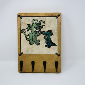 May include: A wooden key holder with a tile featuring a green monster and a green dinosaur. The tile is surrounded by a wooden frame and there are four metal hooks on the bottom.