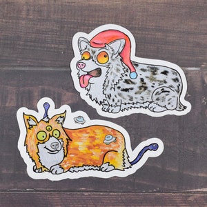 May include: Two illustrated stickers of dogs. One is a grey and white dog wearing a red and white Santa hat. The other is an orange and white dog with three eyes and a blue antenna. Both dogs are on white backgrounds.