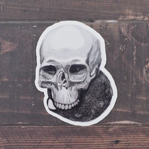 May include: A black and white sticker featuring a detailed illustration of a human skull. The skull is rendered in shades of gray, with visible teeth and eye sockets. The sticker is placed on a wooden surface.