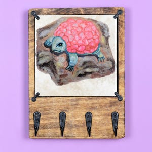 May include: Wooden key holder with three hooks and a painted image of a pink and blue turtle on a rock. The key holder has a dark brown wood finish and black metal hooks.