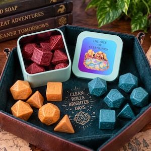 May include: A hexagonal dice tray with dark brown exterior and dark green interior, containing sets of red, orange, and teal polyhedral dice. A light green tin lid features the text THE DICE OF CLEANLINESS and an illustration of a landscape.