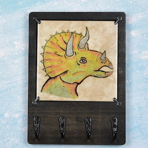 May include: A wooden key holder with a hand-painted triceratops illustration. The dinosaur is yellow, orange, and green. The key holder has four black hooks and a dark brown frame.