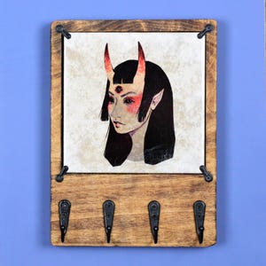 May include: Wooden key holder with three hooks and a framed print of a woman with red horns and a third eye. The print has a white background with a light brown wood frame.