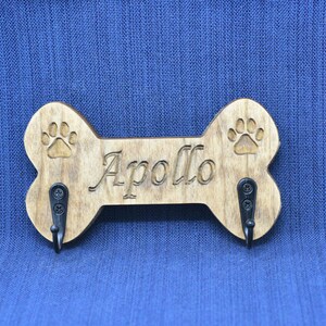 May include: Wooden dog bone shaped key holder with two hooks. The bone is brown and has a paw print design on each end. The name "Apollo" is engraved in the center of the bone.