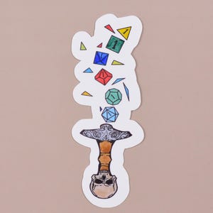 Peut inclure: Un autocollant représentant un crâne transpercé par une épée, avec des dés colorés qui tombent de l'épée. Les dés sont de formes et de tailles variées, avec des chiffres visibles sur certains.
