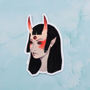 May include: A sticker featuring a stylized illustration of a figure with black hair, red horns, and three eyes. The figure has pale skin with red markings on the cheeks and forehead. The sticker is on a light blue background.