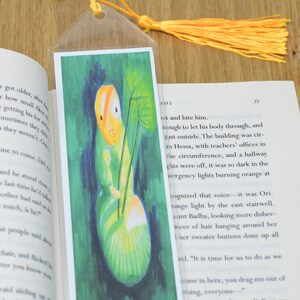 May include: A green and yellow bookmark with a colorful illustration of a caterpillar on a green leaf. The bookmark has a yellow tassel. The text on the bookmark reads: "...enough to let his body through, and just outside. The building was circular in Hessa, with teachers' offices in the circumference, and a hallway..."
