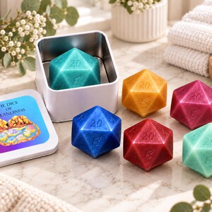 May include: A set of six multi-colored icosahedron-shaped soaps resembling twenty-sided dice. The soaps are teal, yellow, magenta, blue, red, and light green. One soap is inside a white metal tin with a label reading The Dice of Cleanliness.