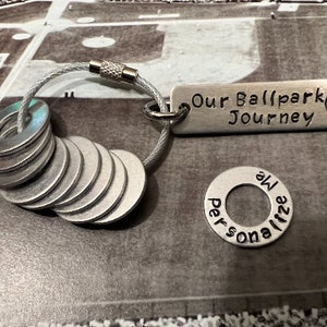 May include: A silver keychain with a baseball-themed charm that reads "Our Ballpark Journey". The charm is attached to a silver chain with a series of silver washers. One of the washers has the words "personalize me" engraved on it.