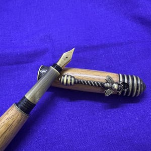 May include: A wooden fountain pen with a gold nib and metal accents. The pen's cap features a decorative bee and honeycomb design. The pen is set against a blue background.
