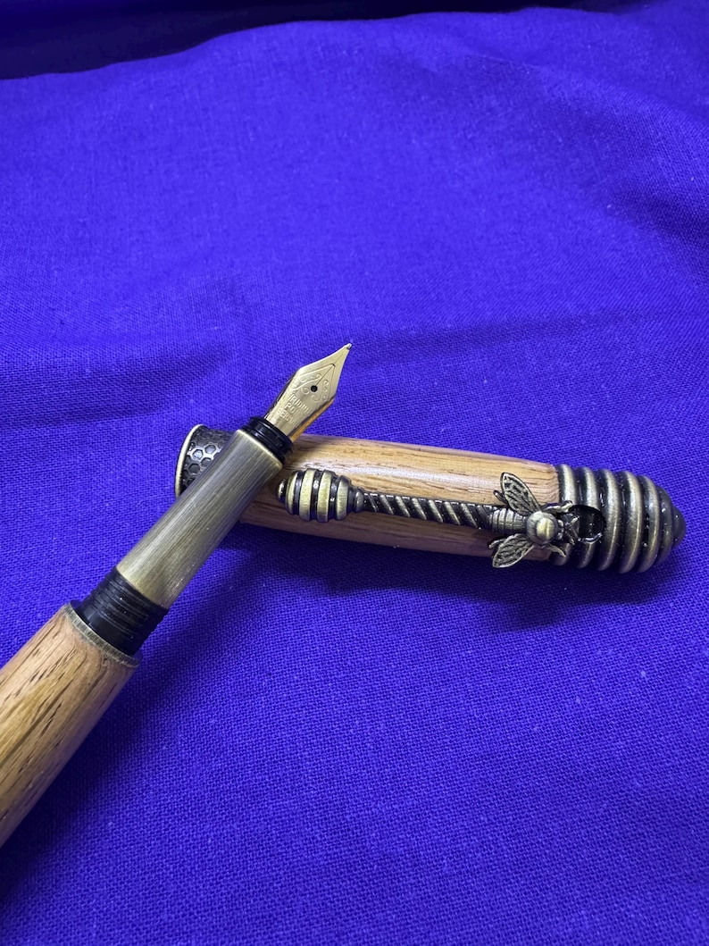 May include: A wooden fountain pen with a gold nib and metal accents. The pen's cap features a decorative bee and honeycomb design. The pen is set against a blue background.