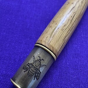 May include: Close-up of a wooden pen with a brass-colored tip. The tip features an engraved bee design. The pen's wooden body has a light brown, textured finish, and the background is a solid blue.