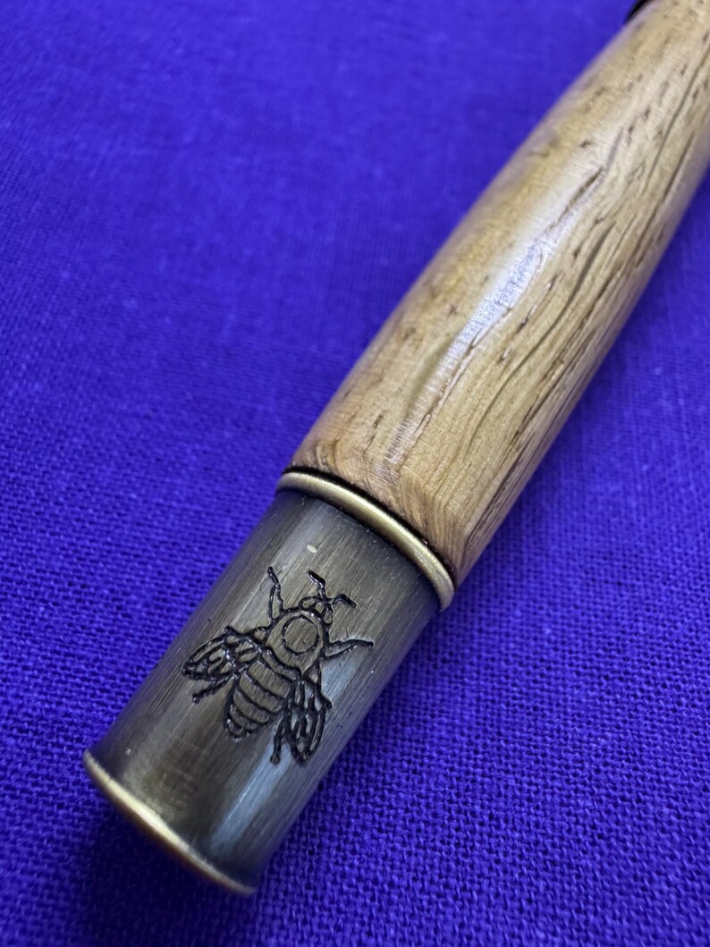 May include: Close-up of a wooden pen with a brass-colored tip. The tip features an engraved bee design. The pen's wooden body has a light brown, textured finish, and the background is a solid blue.