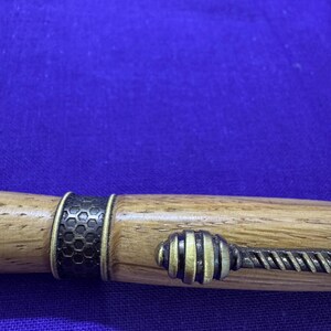 May include: A wooden pen with brass accents. The pen features a dark honeycomb pattern band and a decorative end piece with a twisted design. The pen's body is a light brown color, and the metal accents are gold and black.