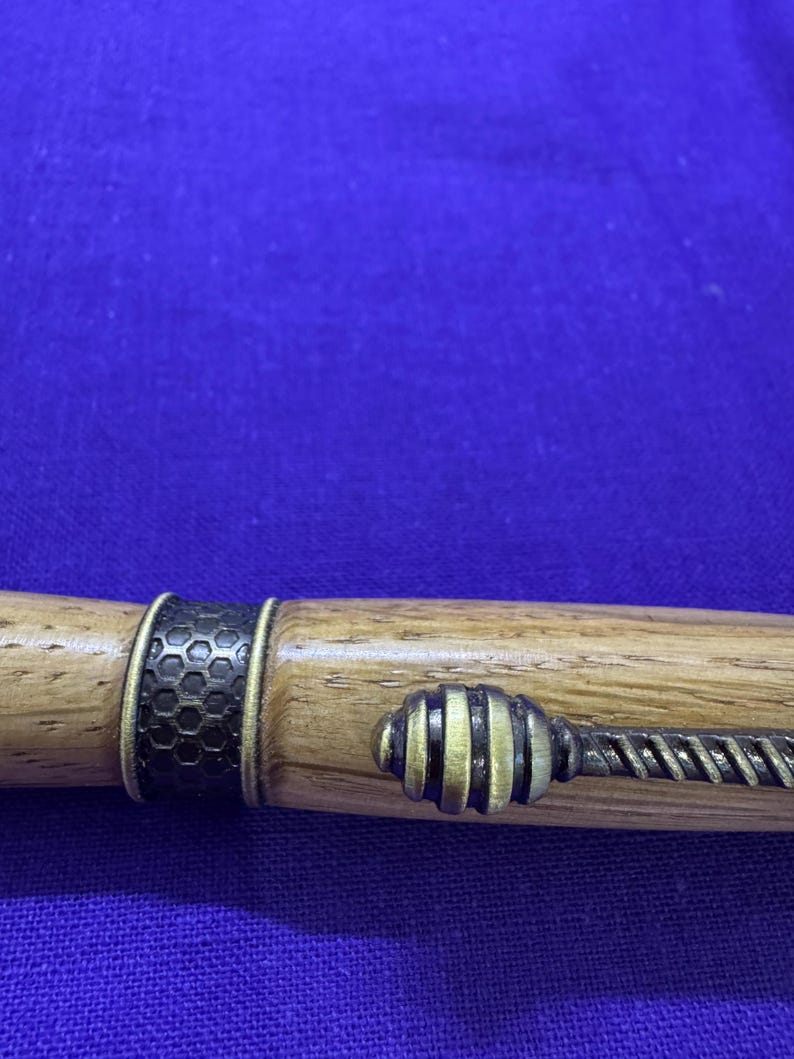May include: A wooden pen with brass accents. The pen features a dark honeycomb pattern band and a decorative end piece with a twisted design. The pen's body is a light brown color, and the metal accents are gold and black.