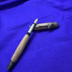 May include: A wooden fountain pen with a gold nib and a decorative bee on the cap. The pen has a light brown barrel and a black and silver accent at the base. The cap is partially open, revealing the nib.