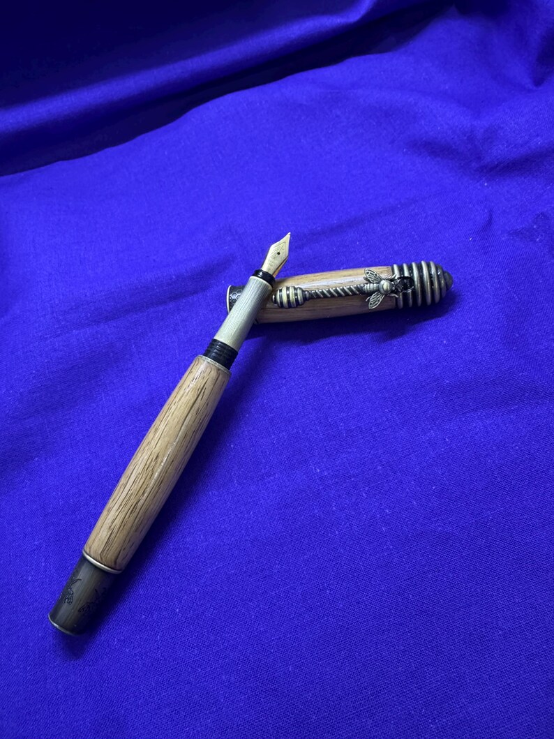 May include: A wooden fountain pen with a gold nib and a decorative bee on the cap. The pen has a light brown barrel and a black and silver accent at the base. The cap is partially open, revealing the nib.