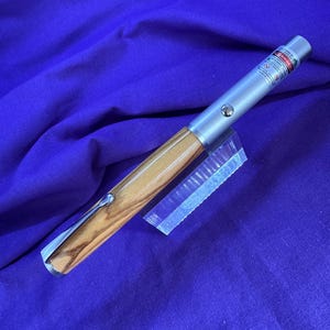 Puede incluir: Un puntero láser plateado y de madera con un soporte de plástico transparente. El puntero láser tiene un mango de madera y un cuerpo de metal plateado. La punta del puntero láser tiene una etiqueta de advertencia roja.