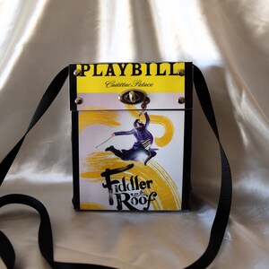 May include: A rectangular crossbody bag featuring the "Fiddler on the Roof" playbill design. The bag has a black strap, a yellow top with the word "PLAYBILL", and a silver clasp. The main image shows a man playing a violin.