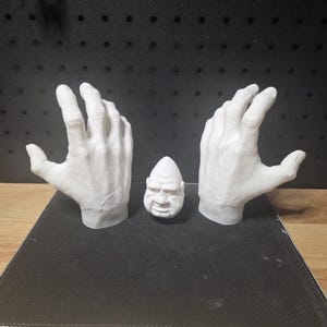 May include: A set of three white 3D printed objects consisting of two realistic human hand sculptures with visible skin texture and veins, positioned on either side of a small, smooth, egg-shaped head figurine with a stern facial expression.