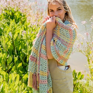 Könnte beinhalten: Eine Frau trägt einen hellgrünen, rosa und gelben Häkel-Schal mit Fransen. Sie steht auf einem Feld mit grünen Pflanzen und Blumen.