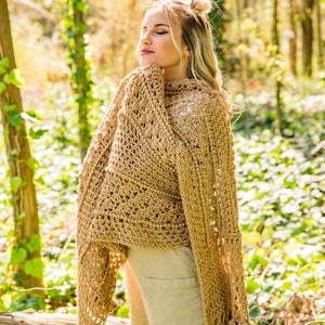 Könnte beinhalten: Eine Frau trägt einen gehäkelten Schal in Beige mit Fransenbesatz. Der Schal hat ein zartes Lochmuster und ist über ihre Schultern gelegt. Sie steht in einer Waldlandschaft mit Bäumen und grünem Laub.