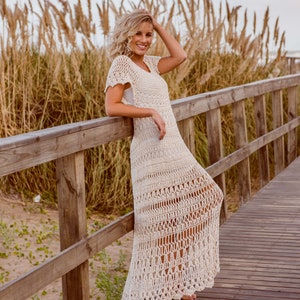 Puede incluir: Una mujer con un vestido largo de crochet blanco se encuentra en un paseo marítimo de madera. Sonríe y tiene la mano en el pelo. El paseo marítimo está rodeado de hierba alta y seca.