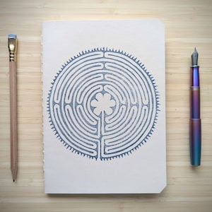 May include: A rectangular notebook with a cream-colored cover featuring a blue circular labyrinth design with a flower at the center. To the left is a wooden pencil with a silver ferrule and eraser, and to the right is a metallic iridescent fountain pen.