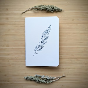 Op de afbeelding: Een wit rechthoekig notitieboek met een donkerblauwe botanische print van een enkel vederachtig blad in het midden van de kaft. Het notitieboek is geplaatst tussen twee gedroogde takjes groen en heeft een strak en eenvoudig ontwerp.