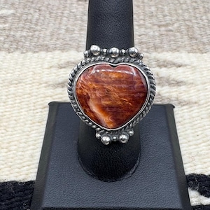 May include: A sterling silver ring featuring a heart-shaped spiny oyster shell. The shell has a warm, reddish-brown hue with intricate patterns. The ring is set on a black display stand against a neutral textile background.