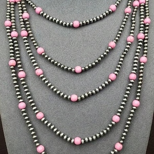 May include: A multi-strand necklace featuring strands of small, silver-colored beads interspersed with larger, round, pink beads. The necklace is displayed against a gray background, showcasing its layered design and color contrast.