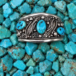 May include: A wide silver cuff bracelet featuring three oval turquoise stones with dark matrix patterns. The metal surface is decorated with intricate leaf motifs, small silver spheres, and twisted wire borders, creating a detailed texture.