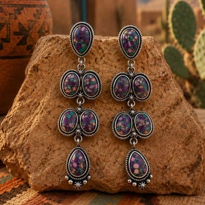 May include: A pair of long dangle earrings featuring multiple oval and teardrop-shaped purple, teal, and tan speckled stones set in silver metal frames with twisted rope detailing and small silver bead accents at the bottom of each earring.