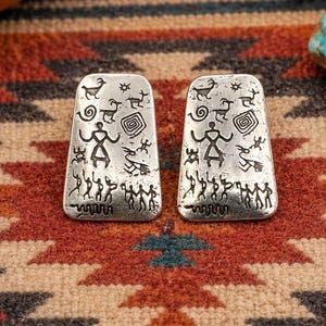 May include: A pair of rectangular silver earrings with rounded corners, featuring etched black petroglyph-style designs of human figures, animals, and geometric shapes across the entire surface of each piece, creating a detailed metallic accessory.