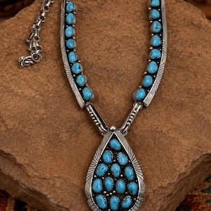 May include: A silver necklace featuring a large teardrop-shaped pendant and two side segments inlaid with multiple small, oval-shaped turquoise stones. The metalwork includes etched line details along the edges and a sturdy silver chain link.