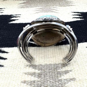 May include: Silver cuff bracelet with a large, oval turquoise stone set in a decorative bezel. The bracelet has a layered design and rests on a neutral-toned patterned textile.